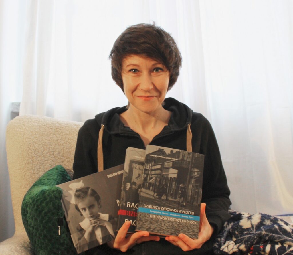 Fotografia przedstawia siedzącą autorkę trzymającą trzy książki swojego autorstwa.